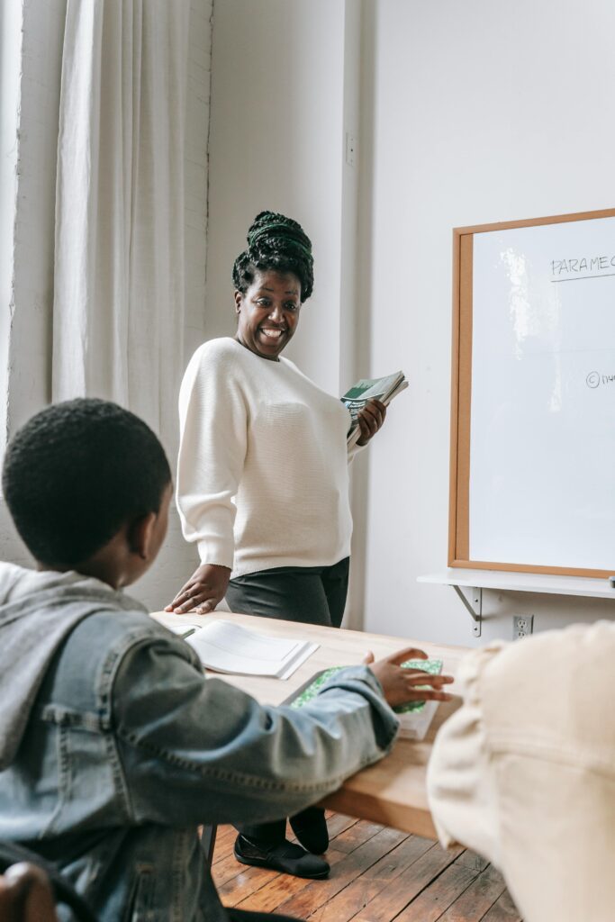 African American female teacher in casual clothes holding books while standing near whiteboard and looking at schoolboy during lesson